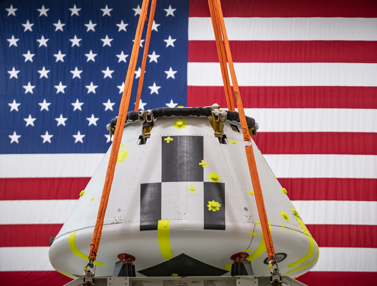Gru solleva una capsula metallica di fattura aeronautica a forma conica mozzata; sul fondo bandiera USA