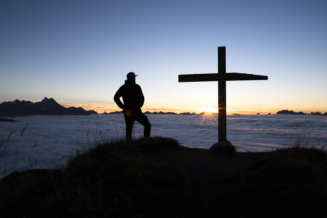 Silhouette of person stands at top of mountain besides cross