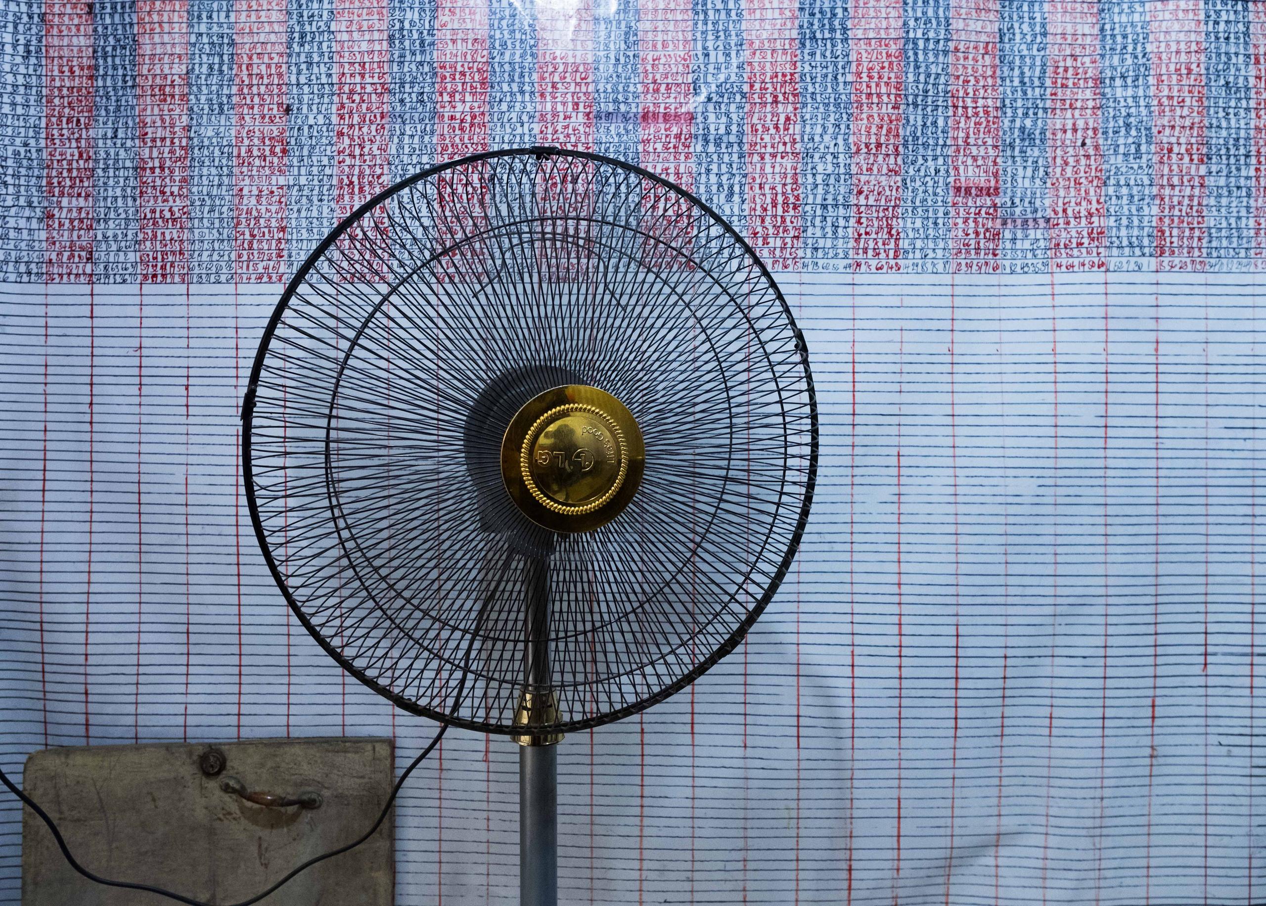 Cabine de loteria. Um ventilador fica em frente aos números da loteria que estão escritos em uma parede.