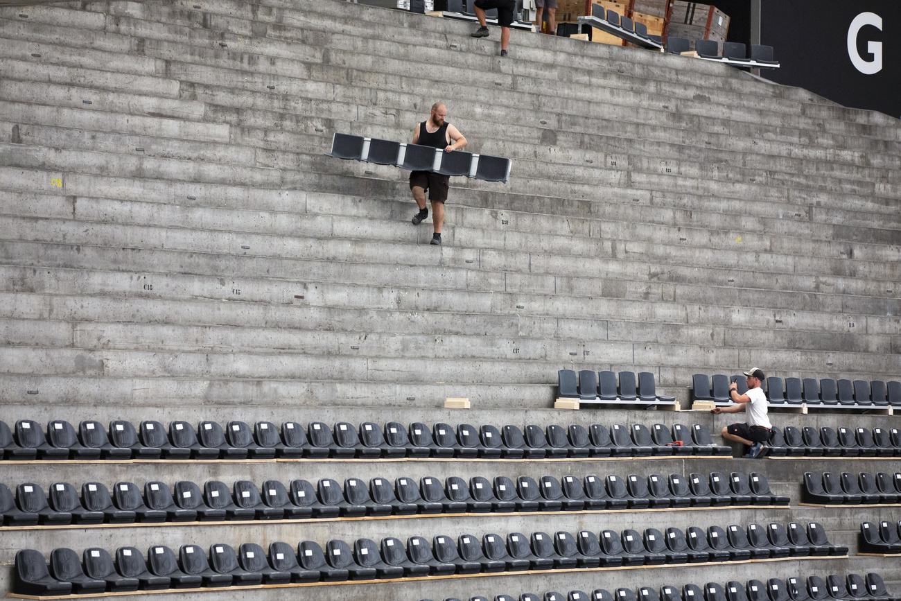 Vista di spalti in cemento armato da stadio sui quali due operai stanno posando file di seggiolini
