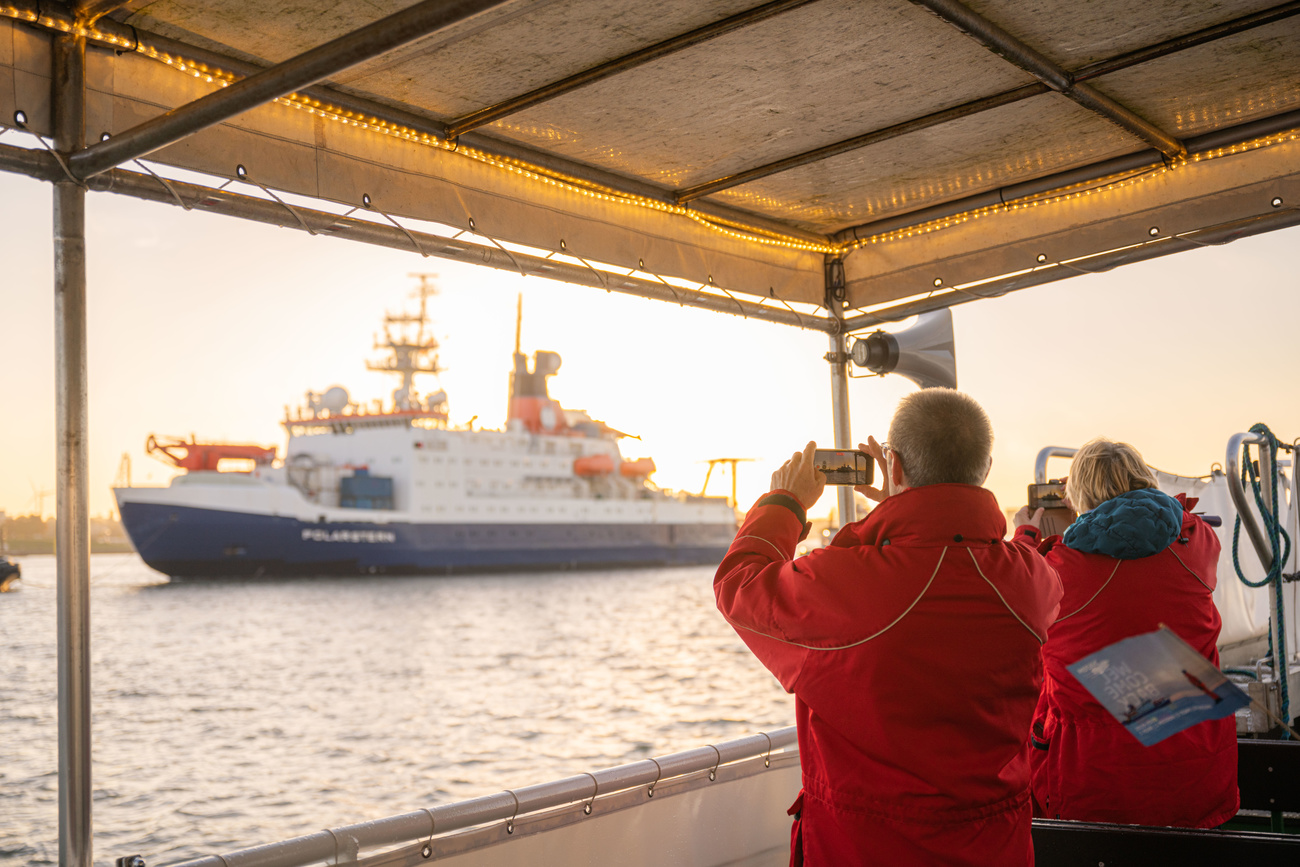 Due persone con giacca a vento fotografano da un molo una nave in arrivo