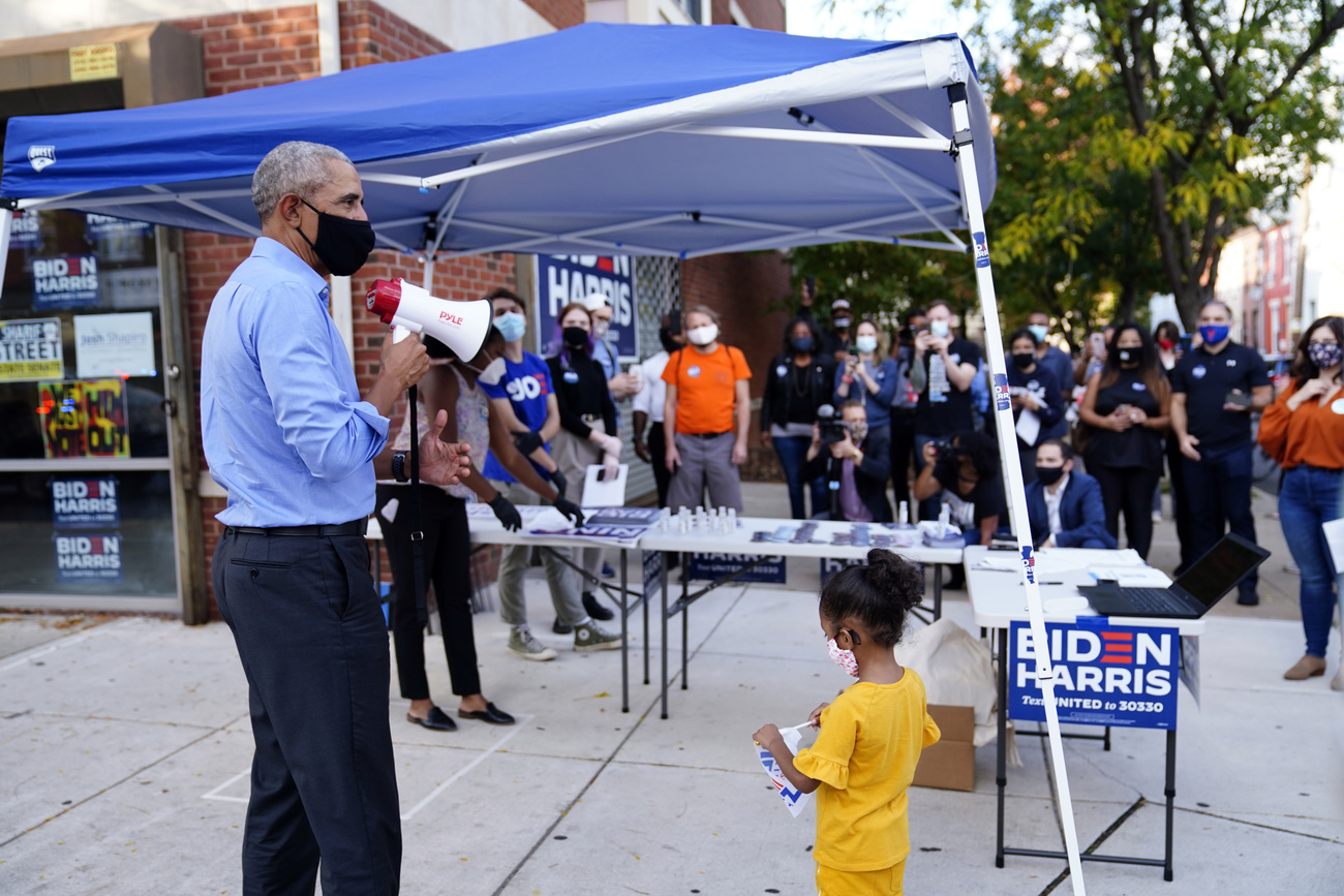 Obama con mascherina e megafono con vicino una bambina nei pressi di un gazebo con scritte Biden/Harris
