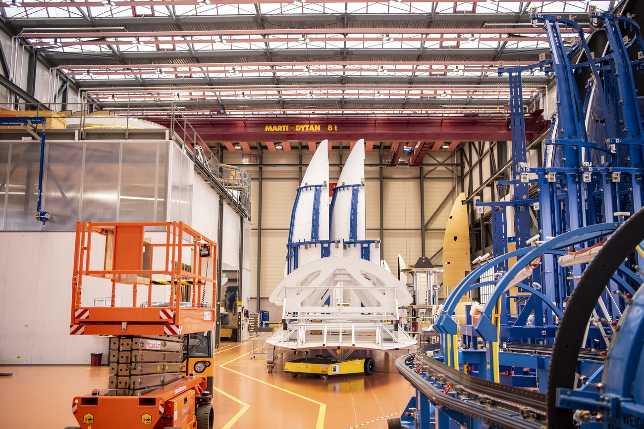 Interno di uno stabilimento con varie macchine e gru, al centro in fondo due carenature a forma di testata di razzo