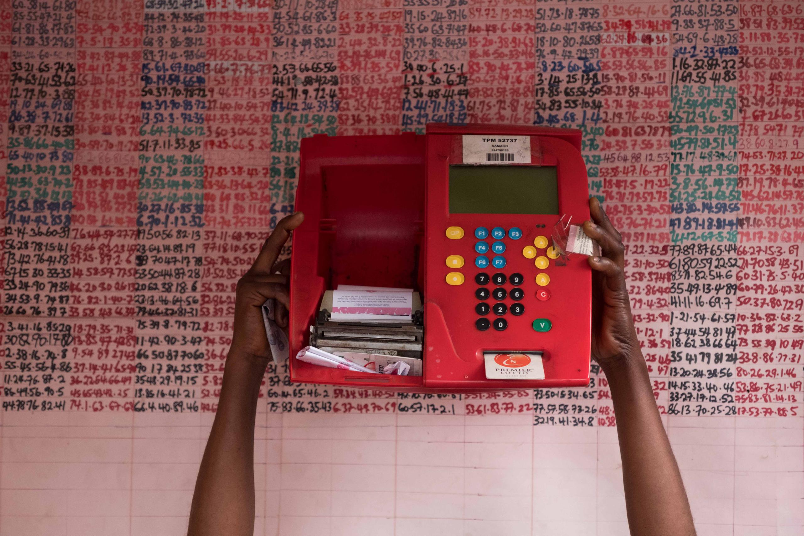 Rudimentary lottery machine held in front of lottery lottery numbers on a wall.