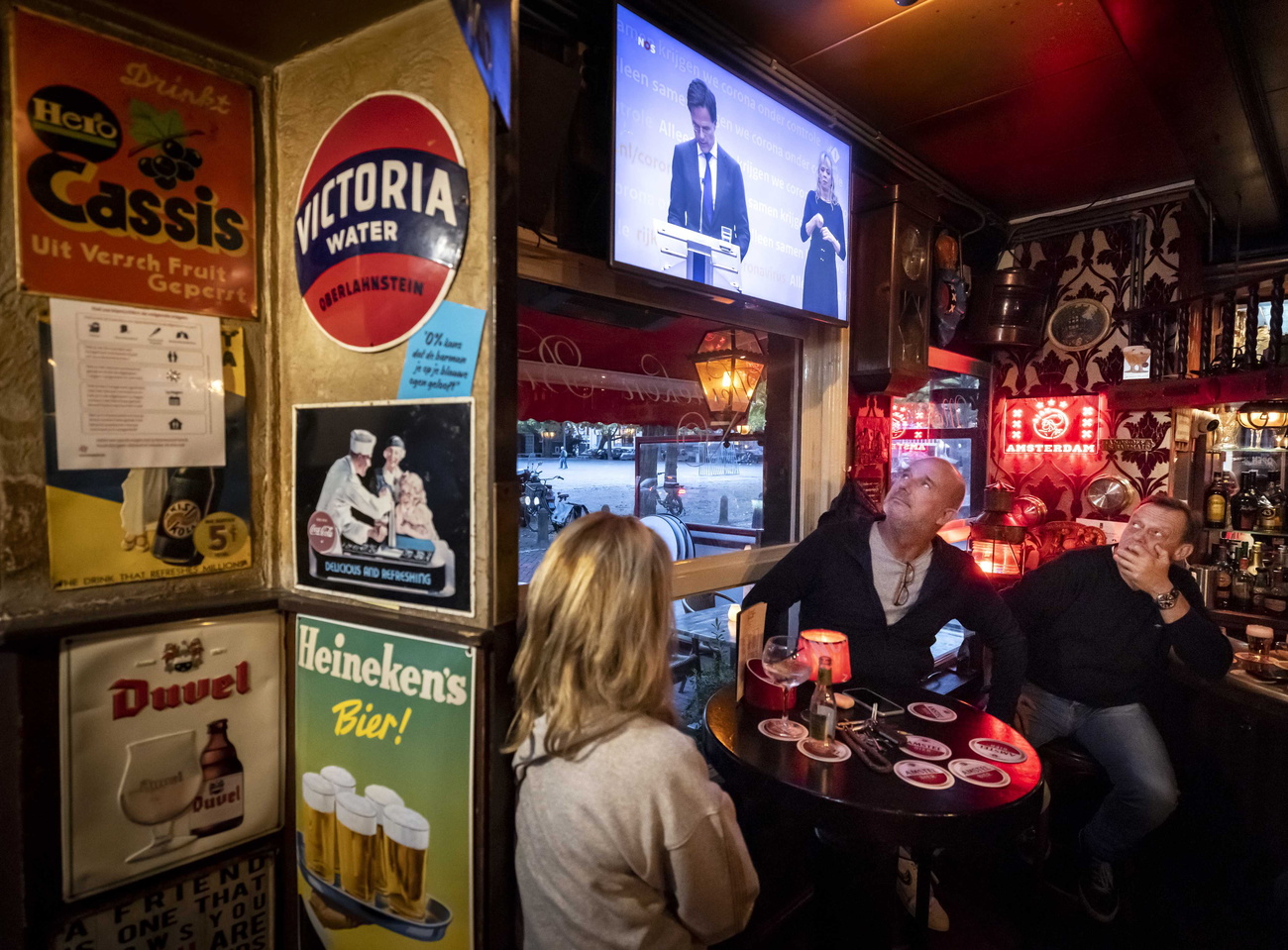 Una donna e due uomini al tavolo di un pub guardano due politici che annunciano qualcosa in tv