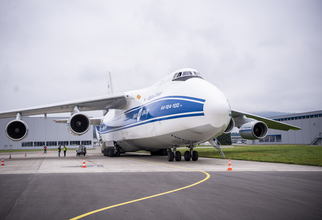 Vista frontale, leggermente obliqua, di aereo con enorme fusoliera in piccolo aeroporto (si intravvedono hangar)