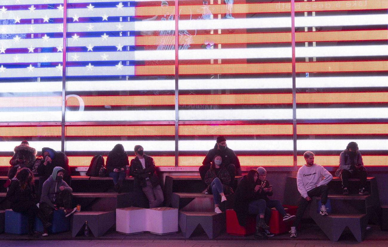 Enorme bandiera statunitense illuminata a led; davanti, persone sedute su subi guardano verso schermi