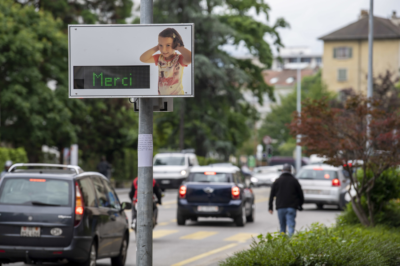 Strada urbana, auto che passano e un cartello con l effige di una bambina che si tappa le orecchie e la scritta luminosa Merci