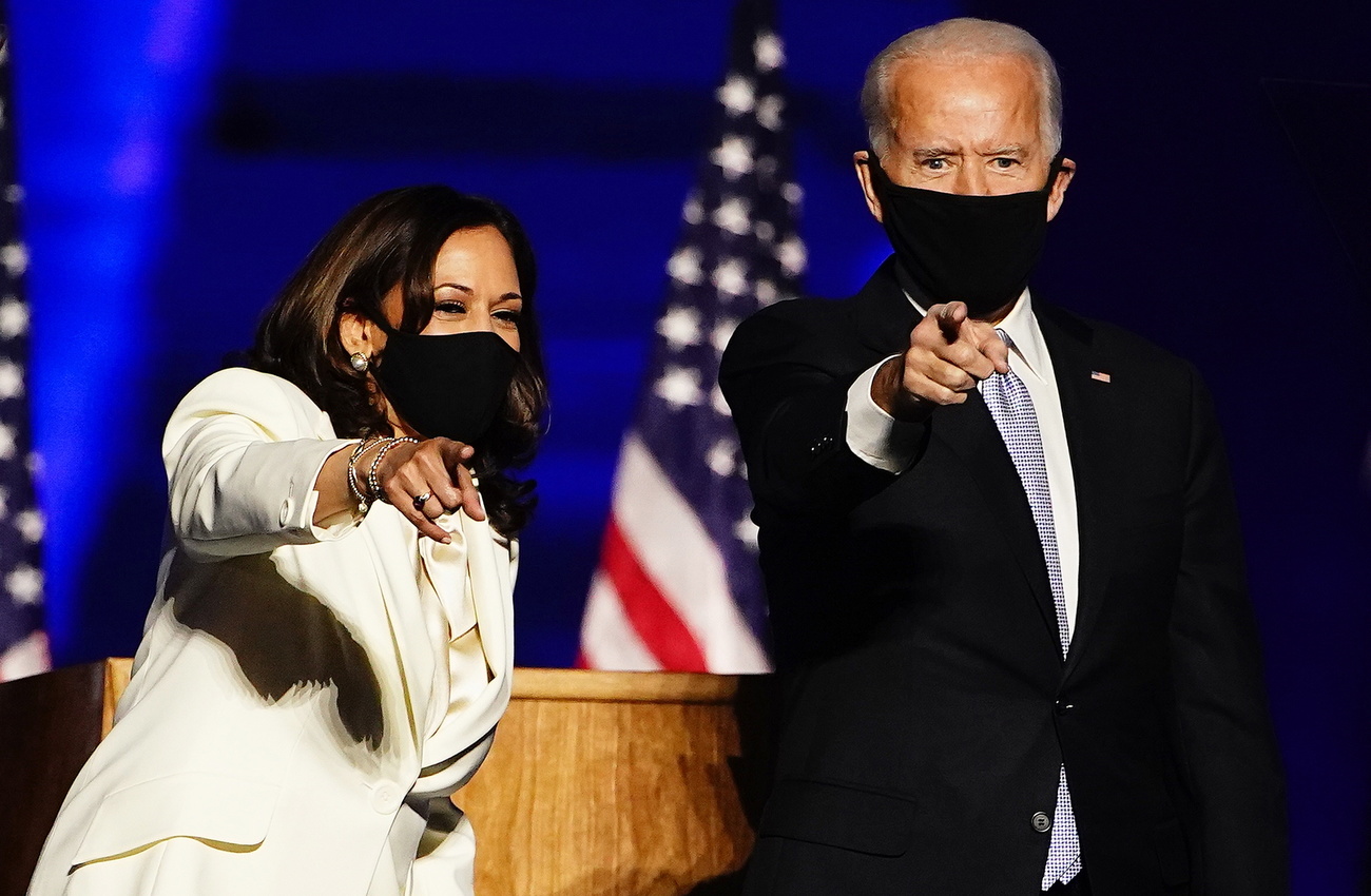 Joe Biden e Kamala Harris con mascherina nera nei pressi di un pulpito e bandiere USA.