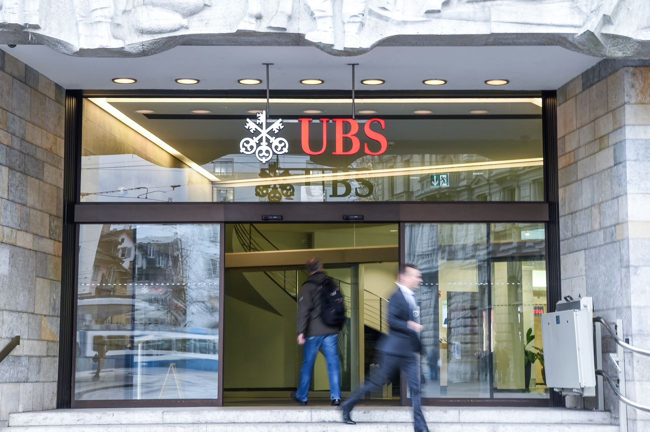 Entrata con porta a vetri di stabile con insegna UBS; un uomo in abiti casual entra, uno in abito formale esce