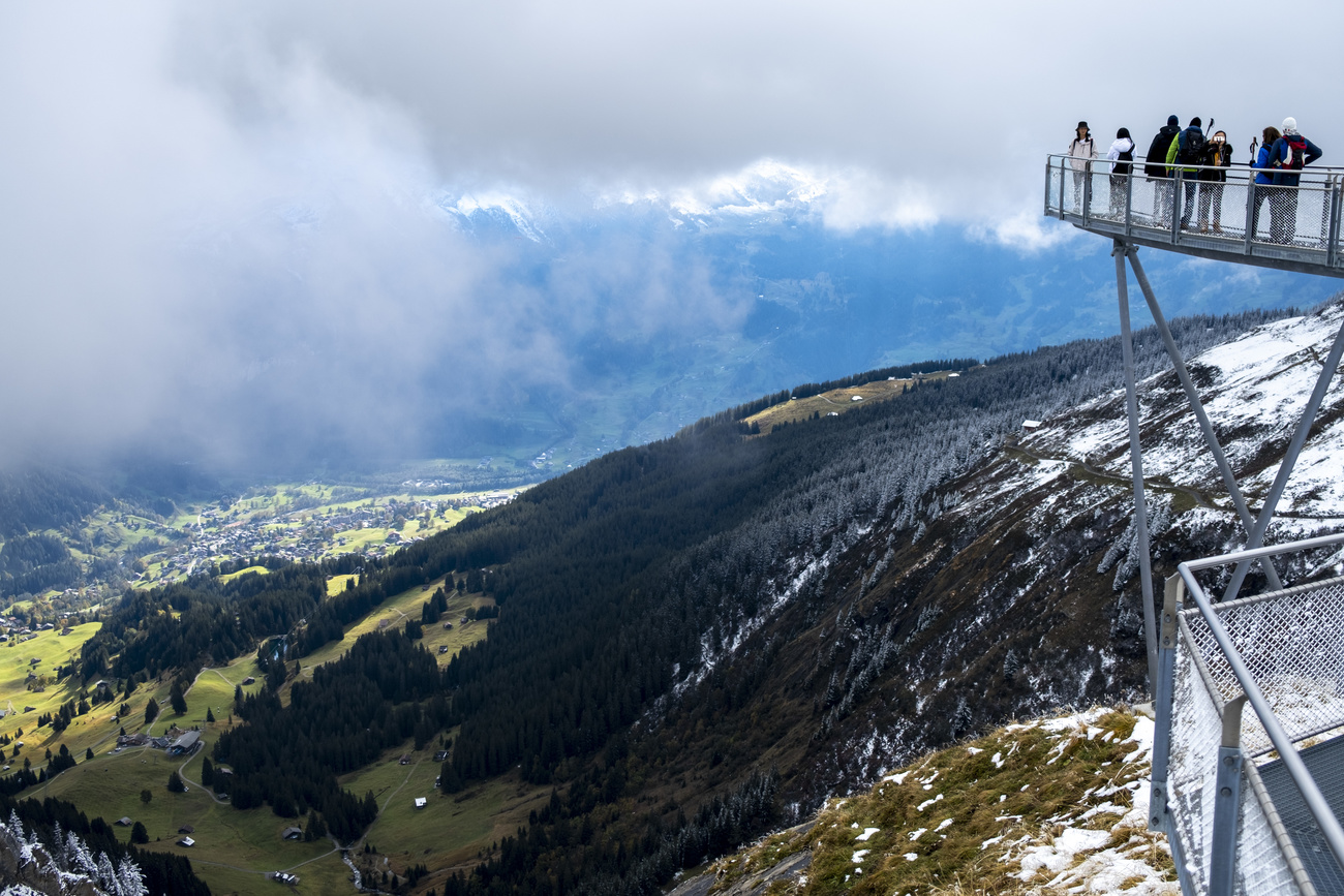 Turisti su una pedana che si sporge da una montagna osservano panorama alpino; tempo nuvoloso
