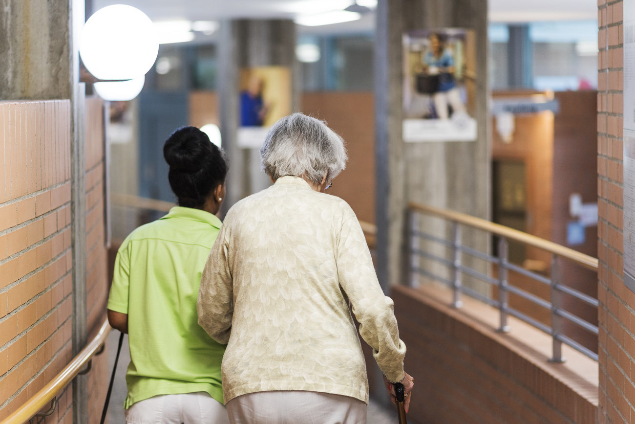 Un assistente di cura accompagna un anziana (di schiena) in una passeggiata in un corridoio di casa anziani
