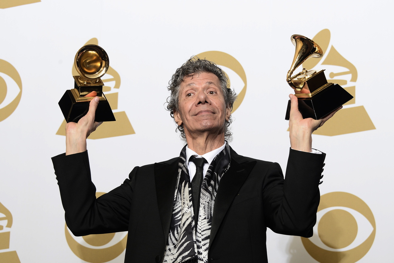 Uomo in cravatta ma casula solleva due Grammy Award mostrando un espressione soddisfatta ai fotografi