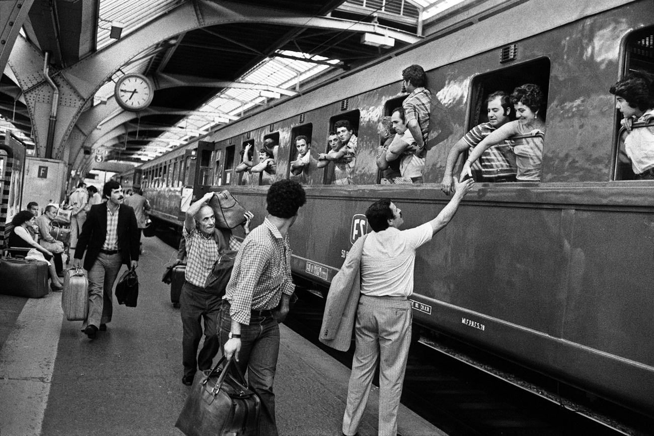 IMmagine in bianco e nero di persone che salutano uomini e coppie sporte dal finestrino di un treno in partenza