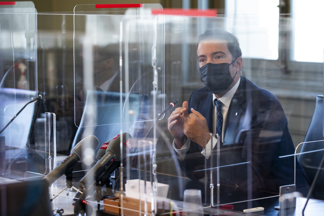 Uomo in abiti formali, visto attraverso diversi pannelli di plexiglas, parla a qualcuno con davanti microfoni di diverse testate