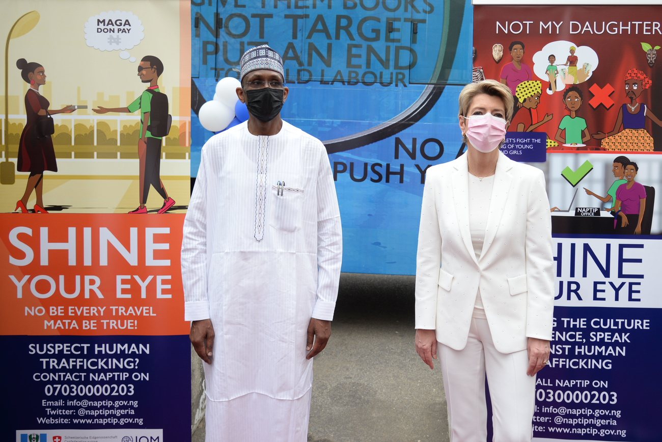 رجل افريقي ووزيرة سويسرية يقفان مرتديين لملابس بيضاء وفي الخلفية لافتات إعلانية