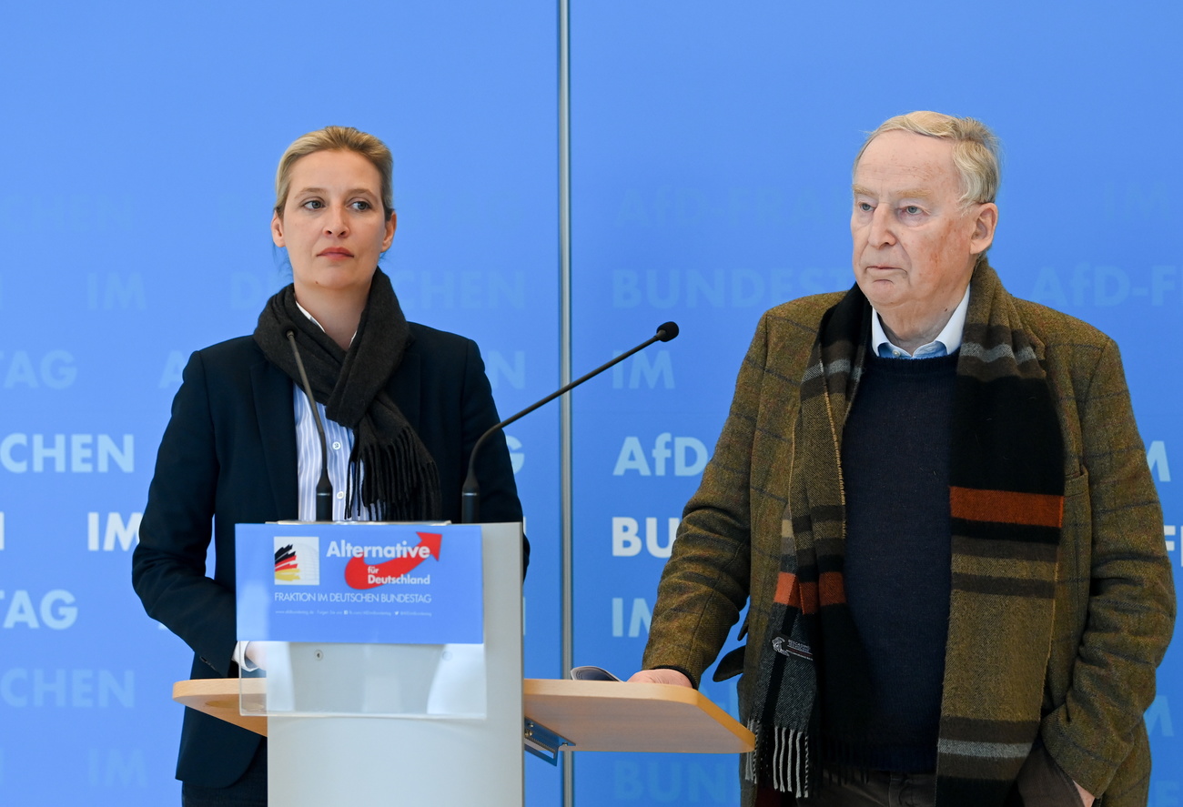 Una donna e un uomo con espressione seria a un pulpito con stemma Alternative für Deutschland, fondo blu