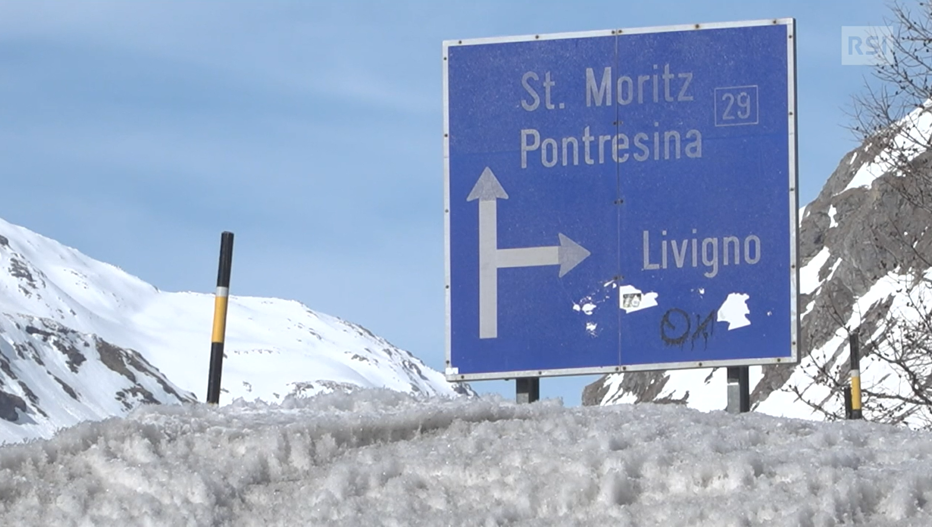 Cartello stradale blu che indica St.Moritz (dritto) e Livigno (destra) in ambiente alpino innevato