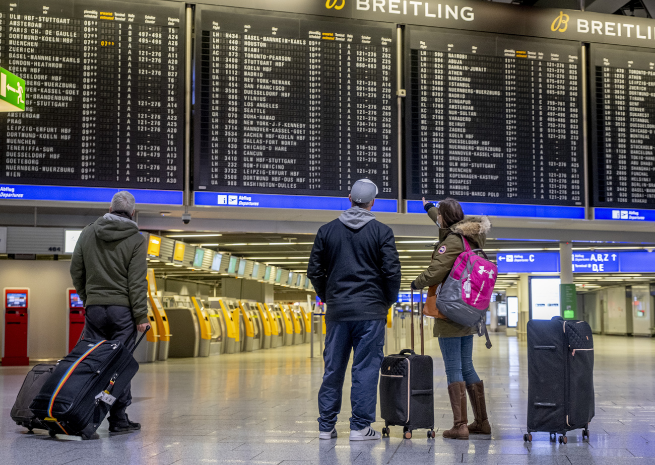 Tre persone in una hall di aeroporto completamente vuota guardano tabellone delle partenze (pieno).
