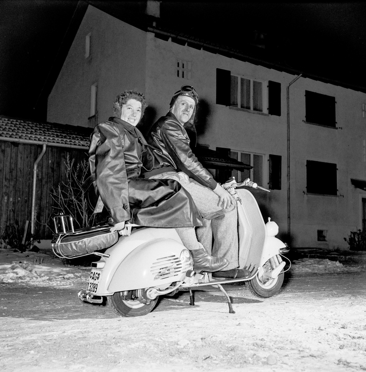 immagine b/n di un uomo e una donna in sella a una vespa fotografati di notte col flash davanti a palazzina di tre piani