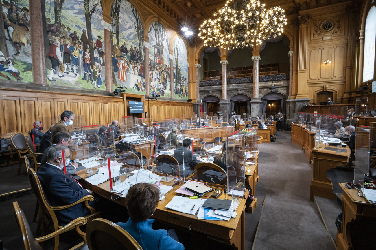 Vista panoramica di sala parlamentare con grande lampadario ed esteso dipinto murale; pannelli in plexiglas sui banchi