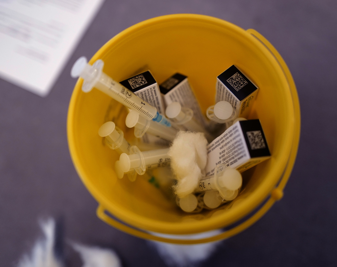 Secchiello giallo da tavolo contenente siringhe e confezioni di un farmaco.