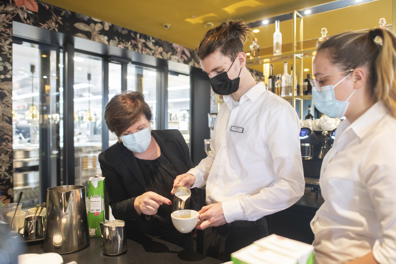 Un giovane cameriere prepara il cappuccino mentre una collega più anziana lo osserva e corregge.