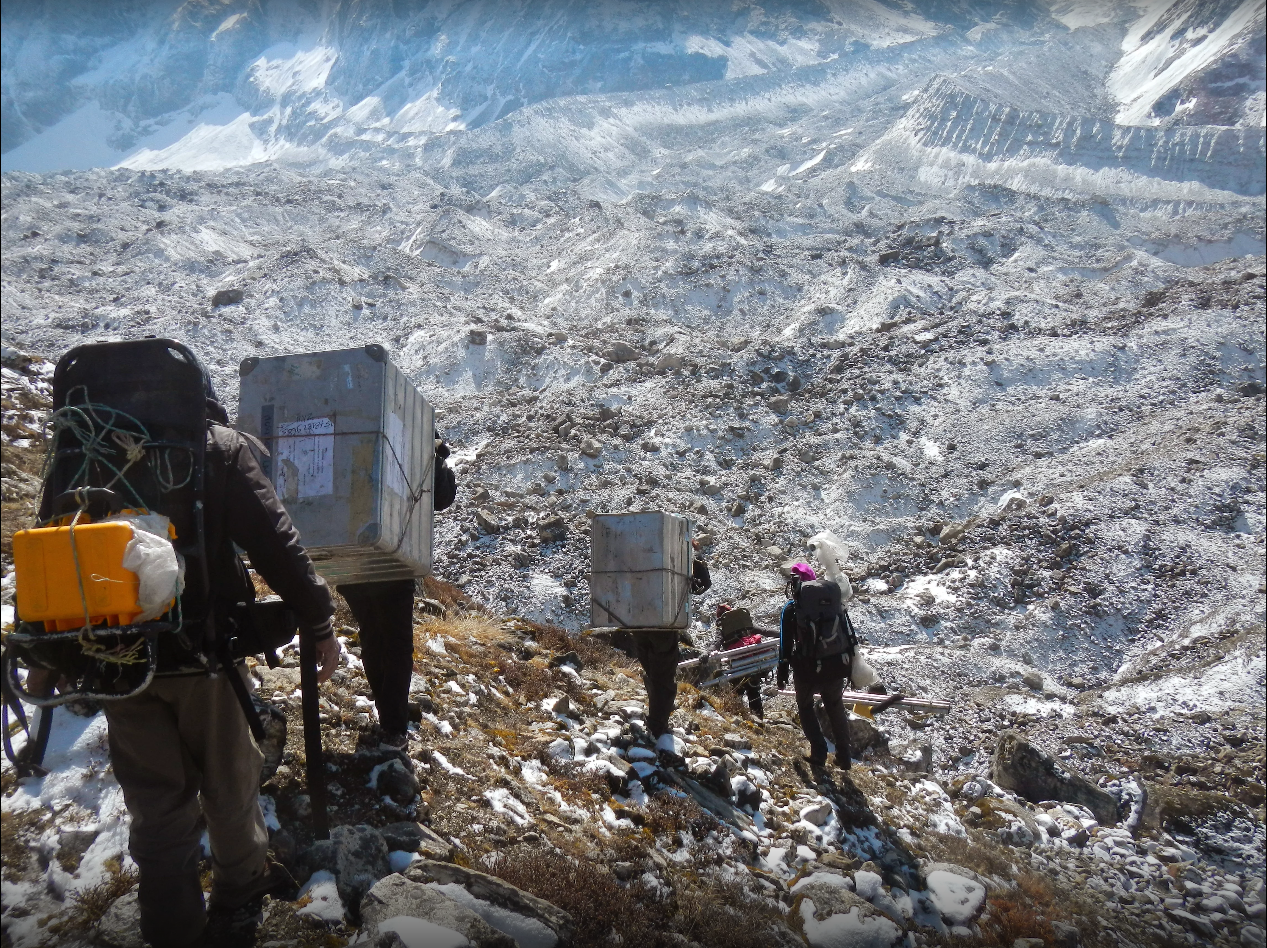 gente che cammina su un ghiacciaio e trasporta in spalla grosse casse di metallo