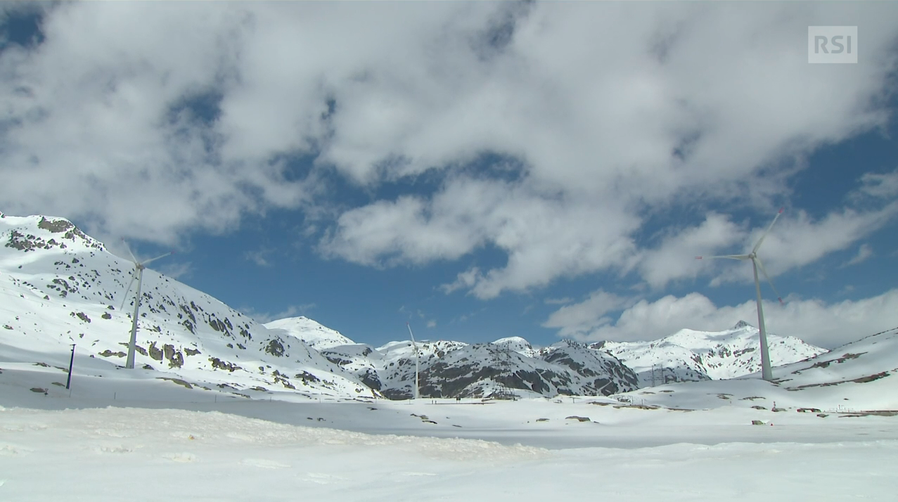 Turbine eoliche in ambiente alpino aperto e innevato in giorno nuvoloso.