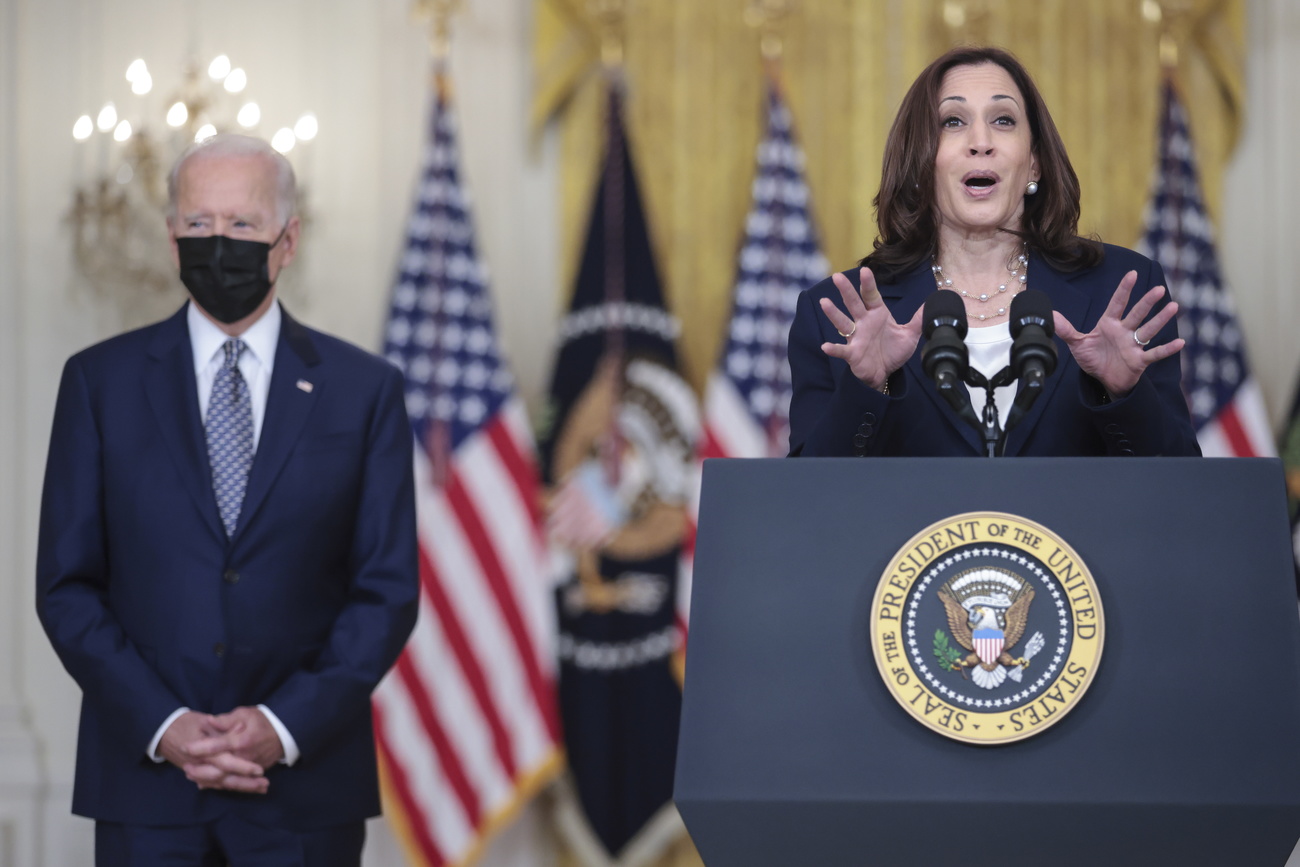 Kamala Harris in primo piano parla davanti a un microfono. Dietro di lei Joe Biden con la mascherina nera