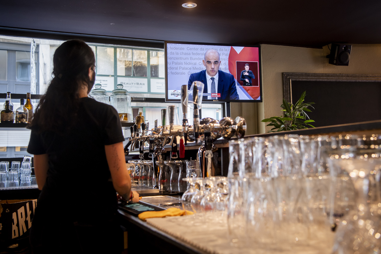 Una barwoman segue in tv il discorso di Alain Berset da dietro un bancone.