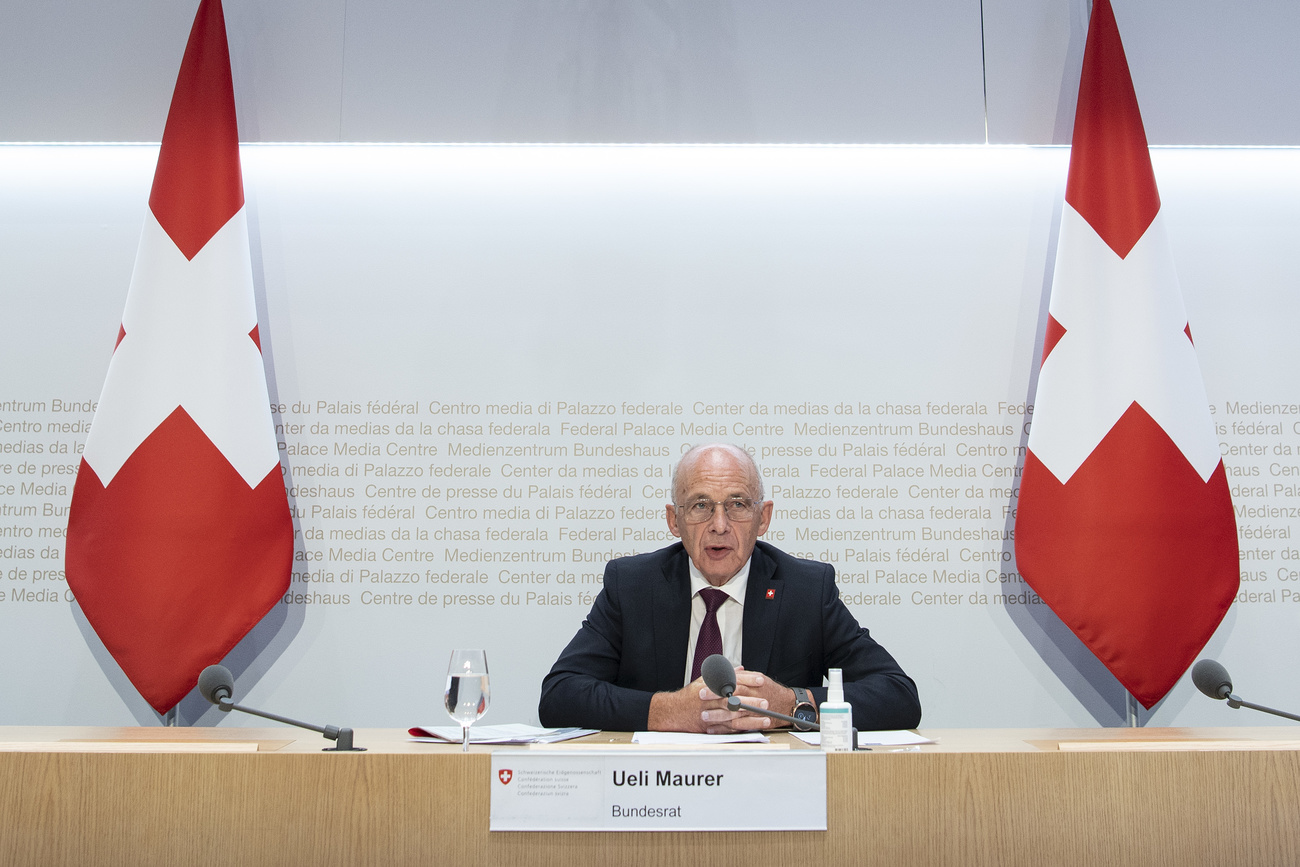 رجل يتحدث وهو جالس أمام مصدح متوسطا علمين سويسريين