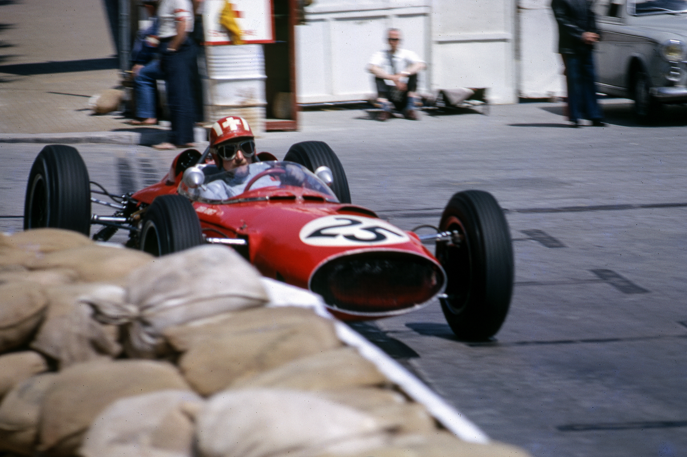 Voiture de course des années 1960 avec un pilote portant un casque rouge à croix blanche.