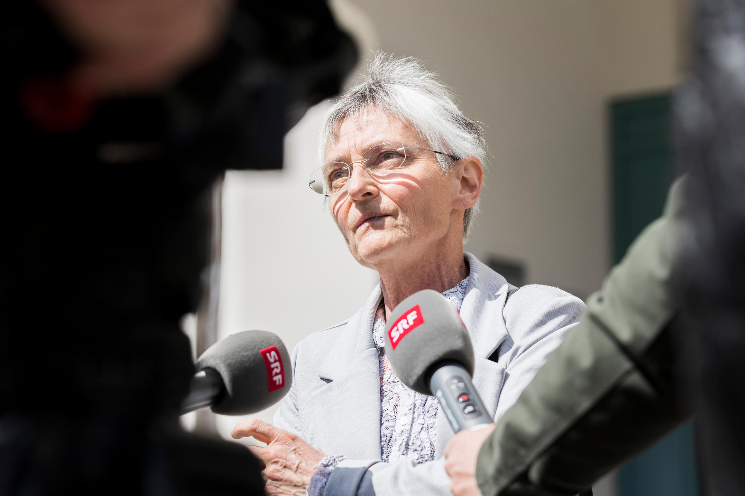 Donna con capelli bianchi corti risponde a domande di giornalisti radio-tv (microfoni con logo SRF)