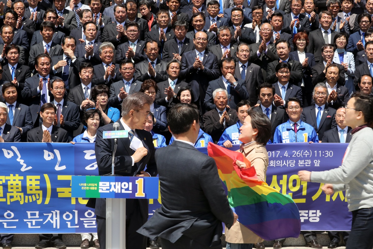 attivista con bandiera arcobaleno si avvicina a un uomo che sta parlando davanti a un gruppo di persone