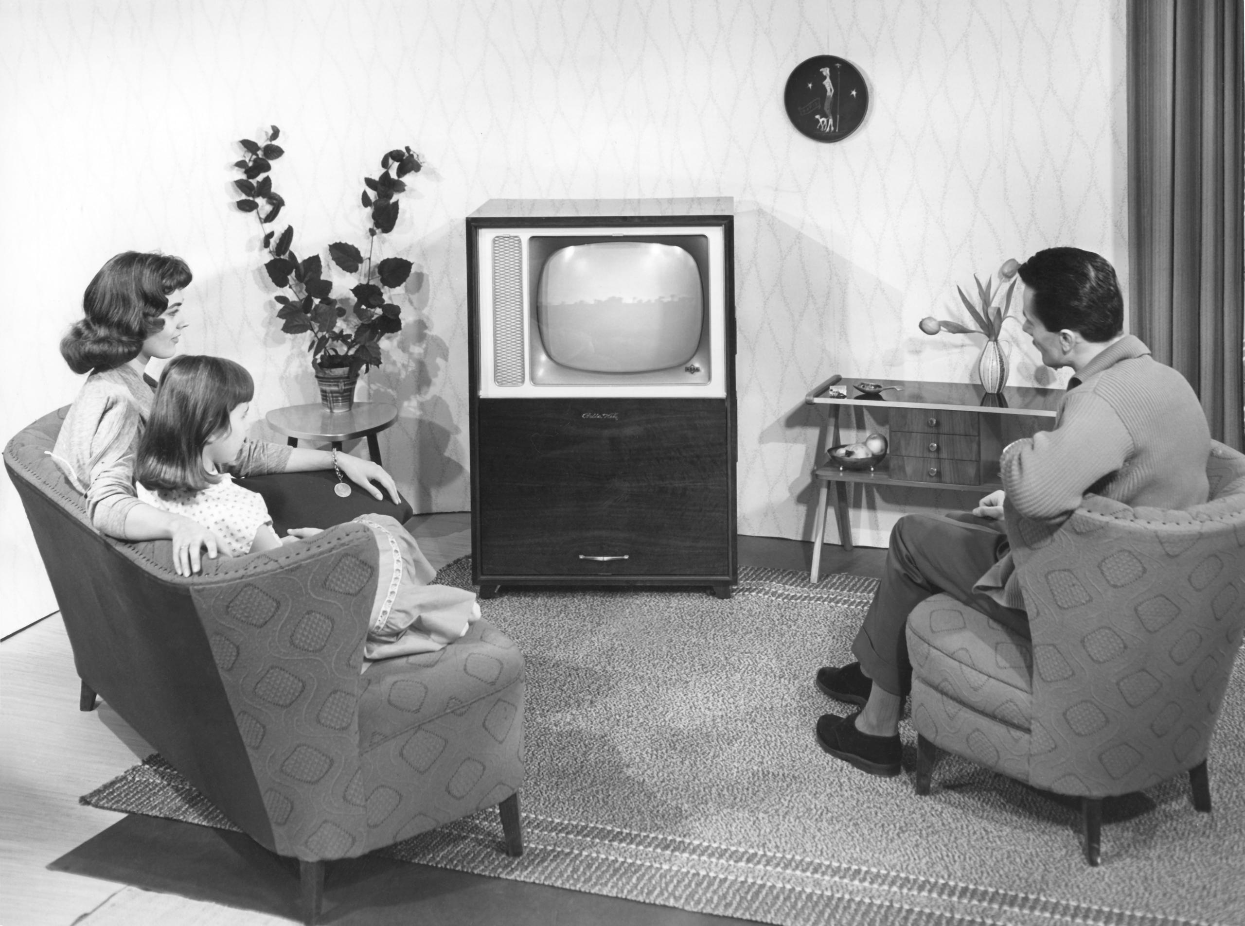 Foto in b/n di famiglia in soggiorno anni 60, con padre, madre e figlia seduti sul divano e rivolti verso il televisore