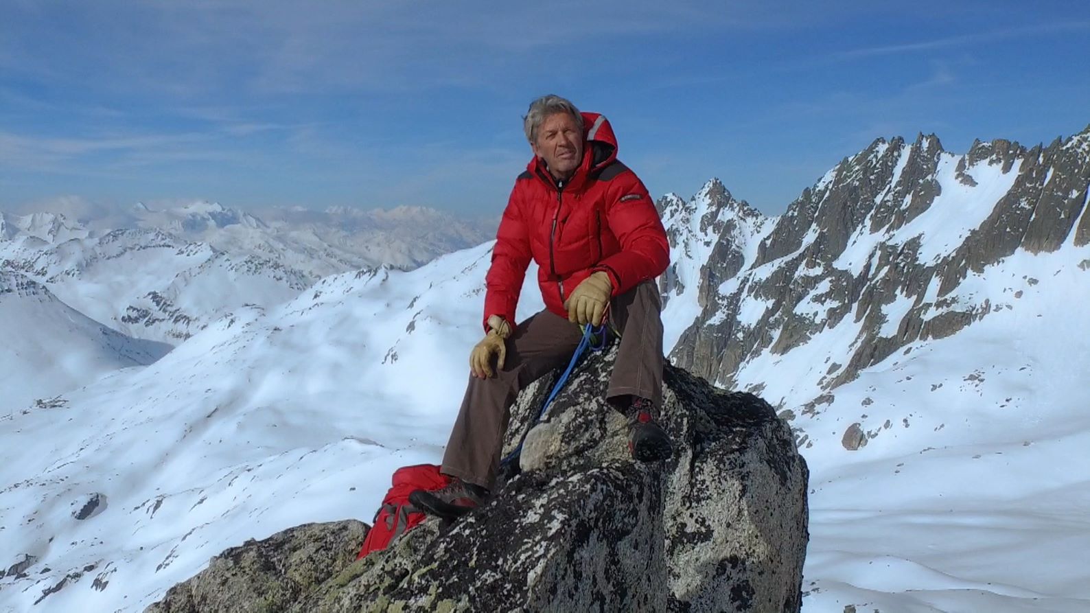 Mann in roter Jacke, der in verschneiten Bergen auf einem Felsen sitzt