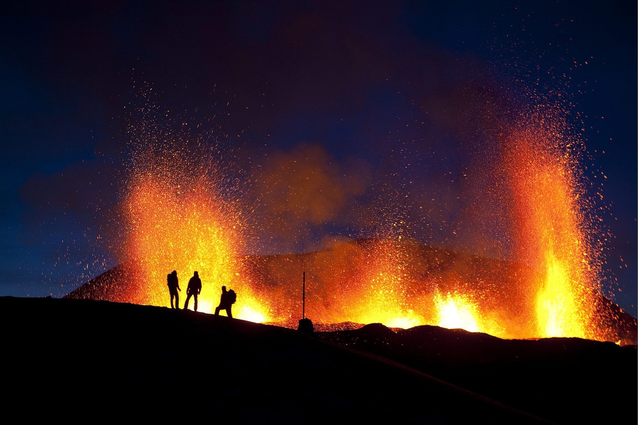 Un volcán en erupción con la silueta de unos hombres en la cresta del propio volcán