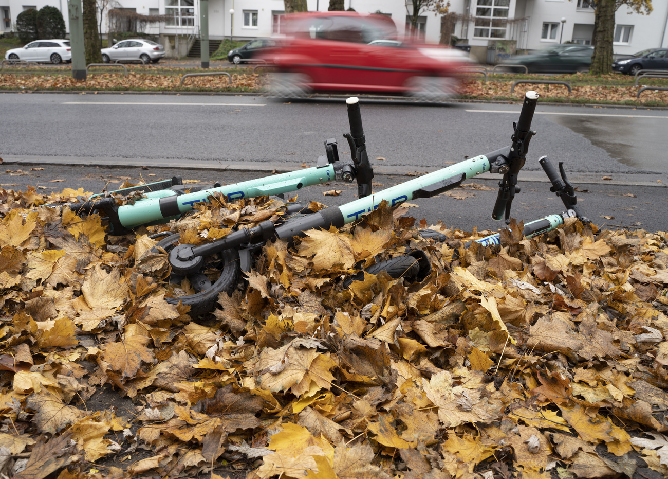 E-scooter lying in leaves at the side of a road with a car passing in the background.