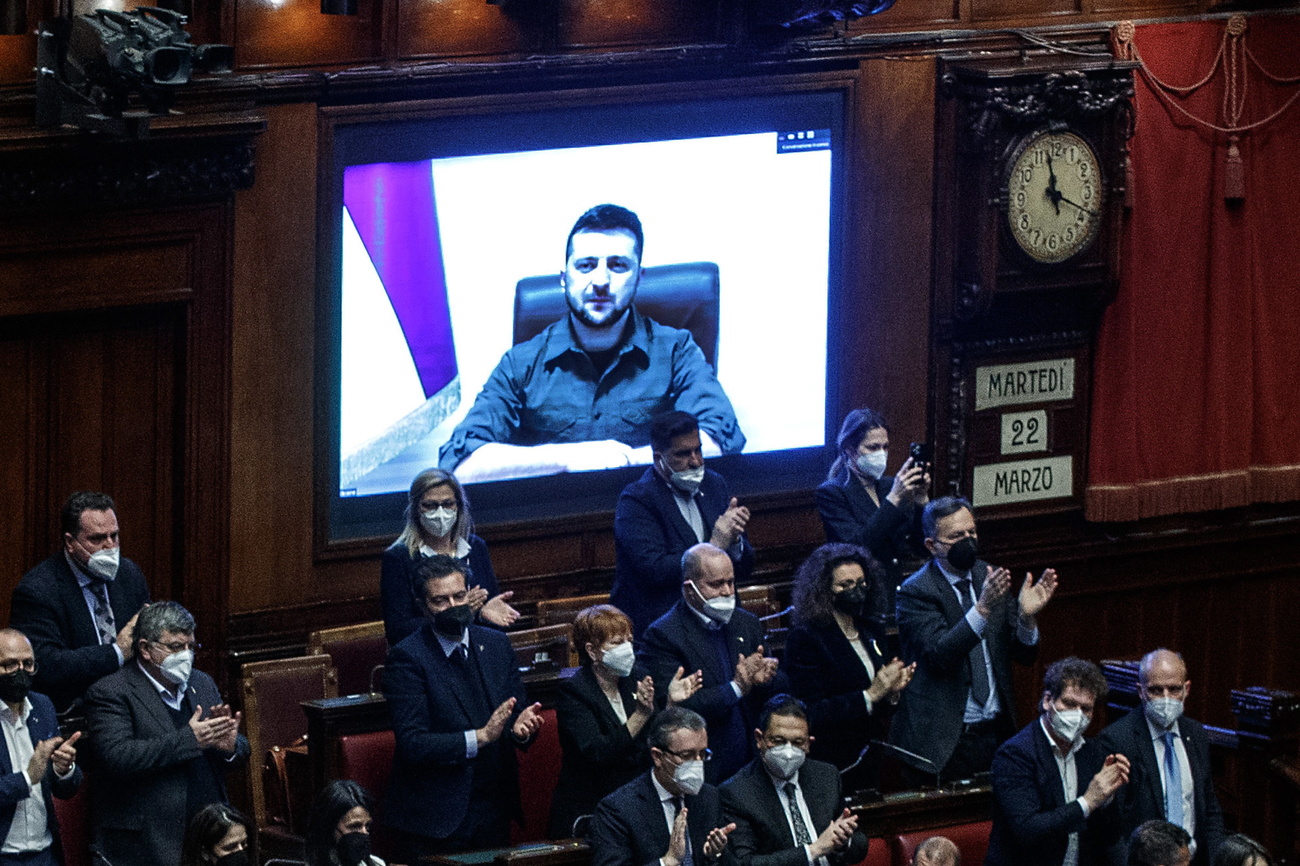 persone in piedi applaudono. Dietro di loro su uno schermo apapre un uomo, seduto a una scrivania