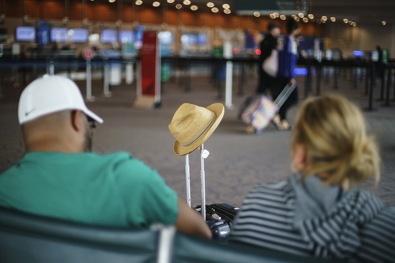 uomo e donna seduti su poltrone in aeroporto fotografati di spalle. davanti a loro un trolley