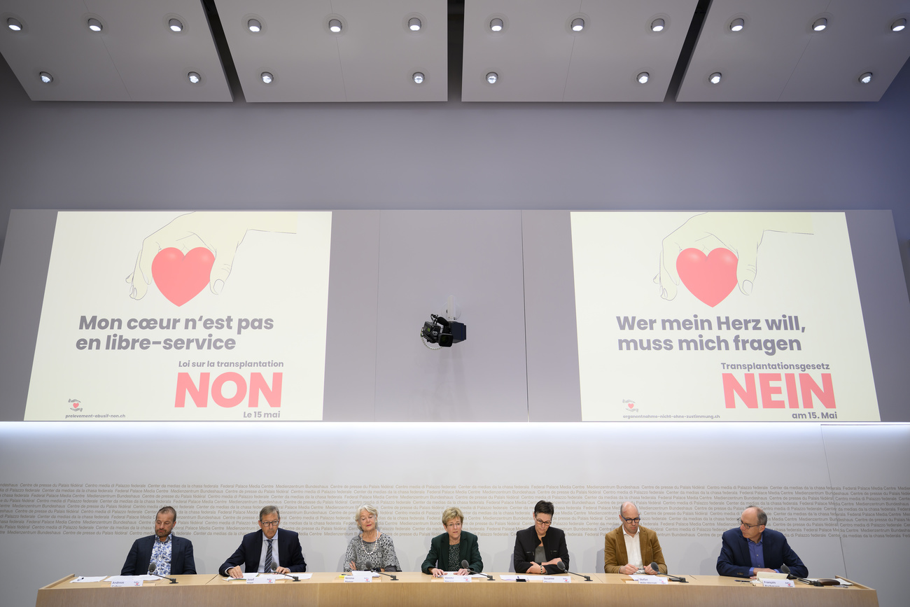 7 persone sedute a un tavolo, dietro di loro due cartelli con disegno di cuore e la parola no in francese e in tedesco