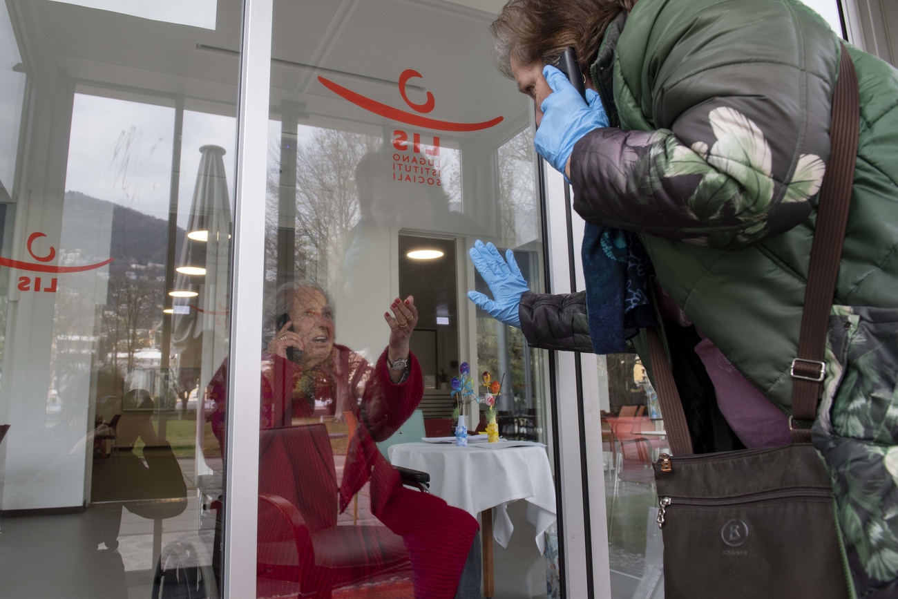 Mulher idosa atrás da porta de vidro fechada falando ao telefone com uma pessoa esperando do lado de fora