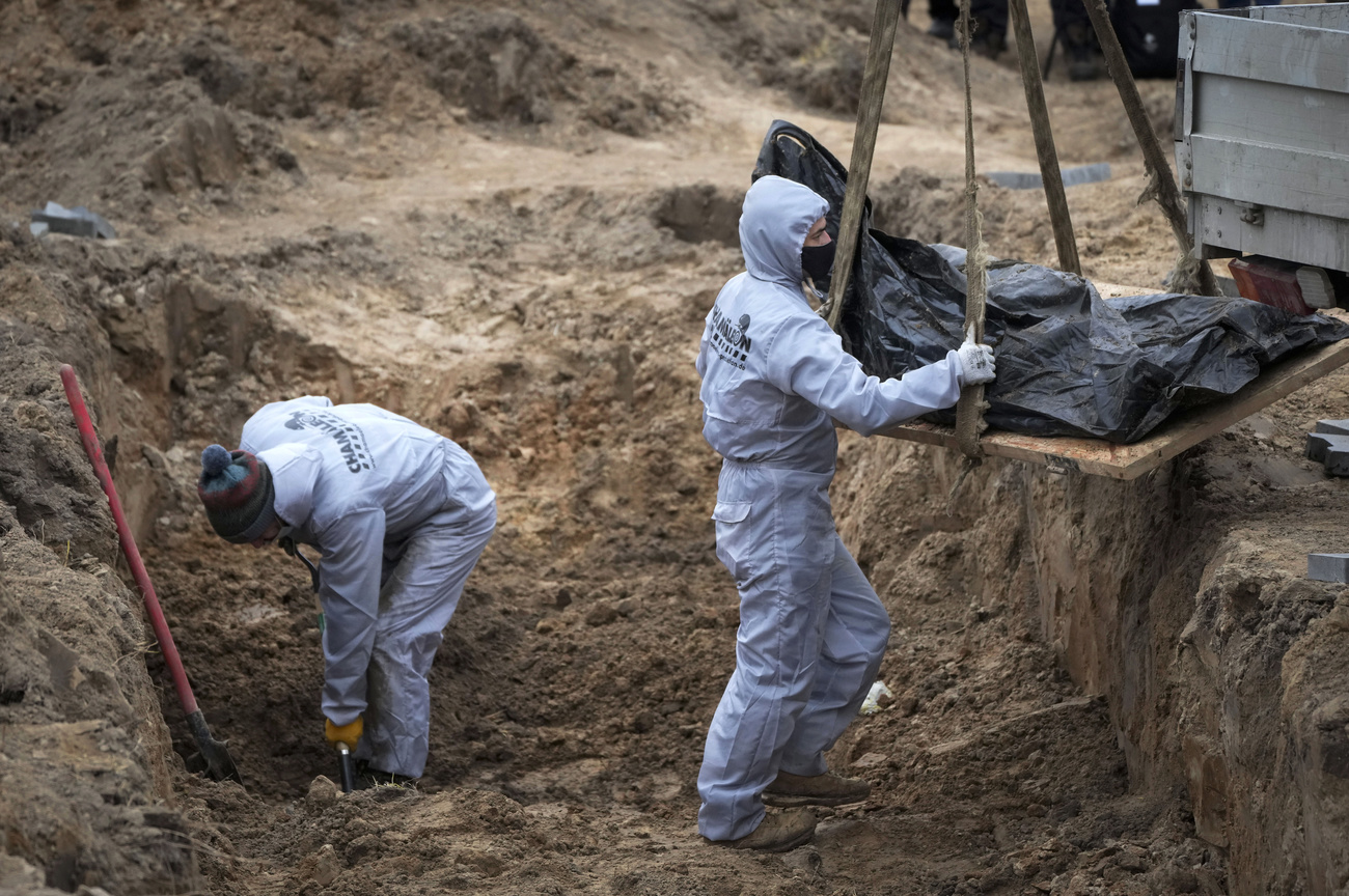 uomo in tenuta protettiva solleva, avvolto in un sacco nero, un corpo estratto da una fossa comune