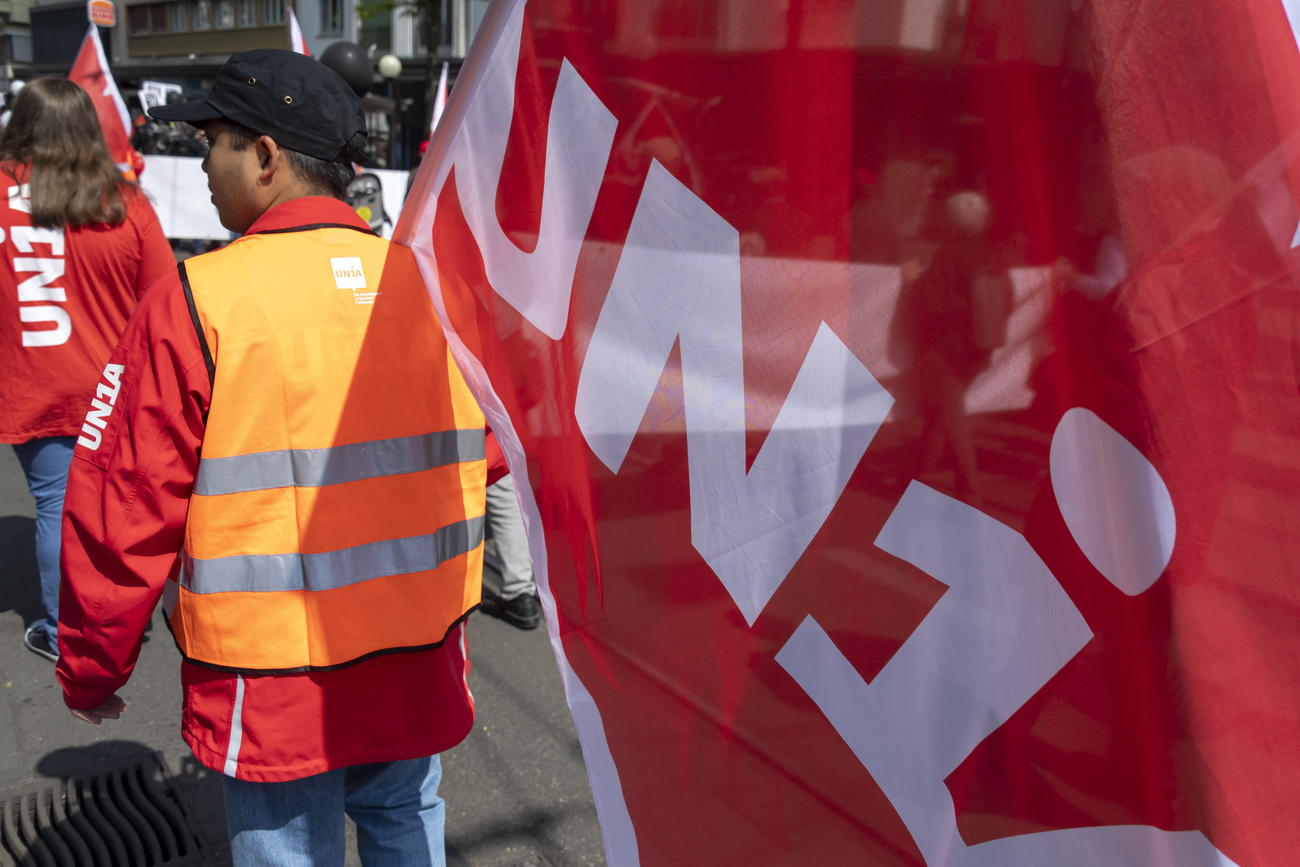 uomo con gilet di sicurezza arancione fotografato di spalle tiene una bandiera di unia (scritta bianca su sfondo rosso)