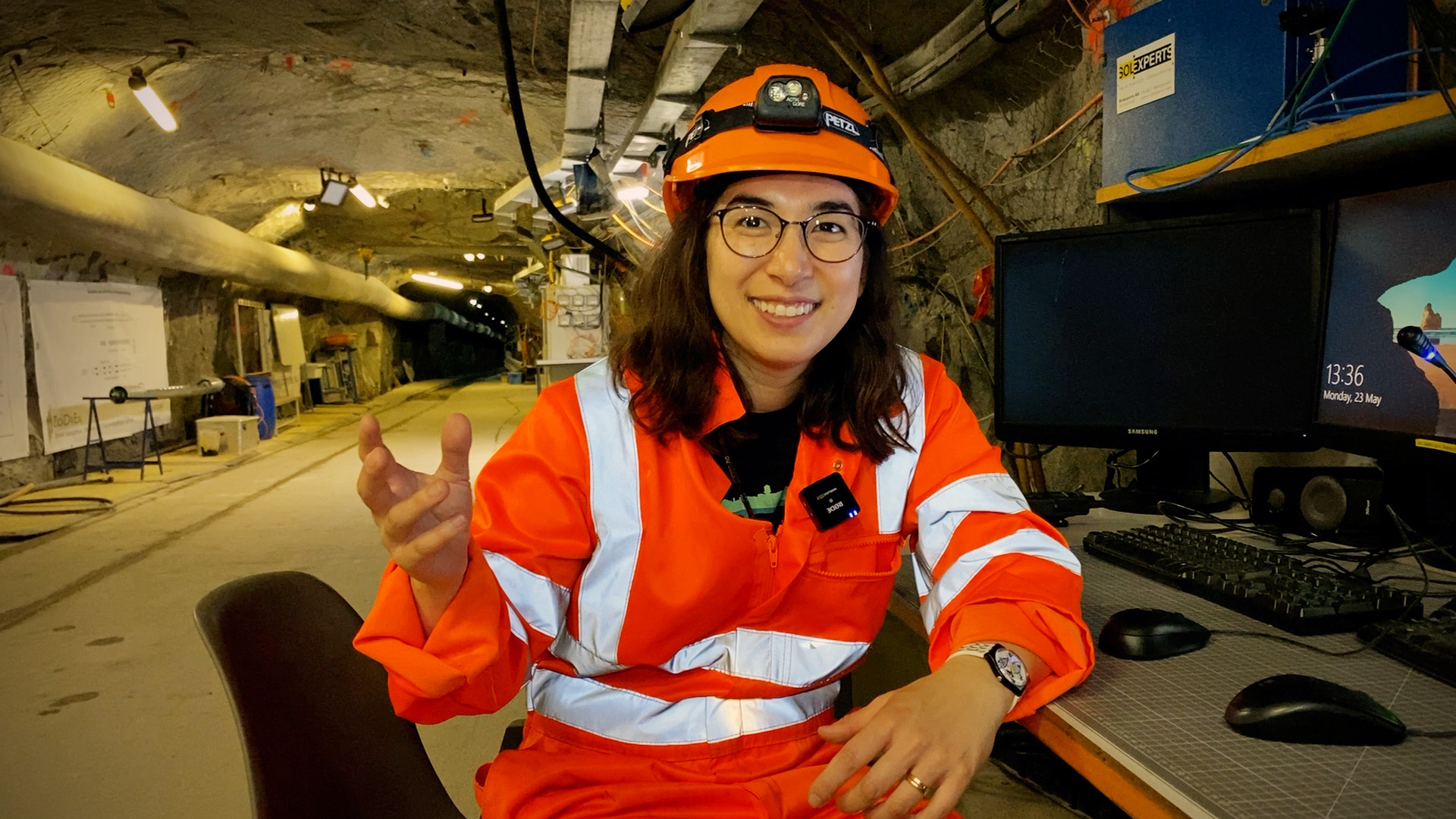 Junge Frau in orangem Bauarbeitergewand sitzt auf einem Stuhl in einem unverputzten Tunnel