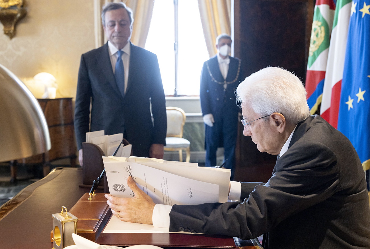 mattarella seduto alla sua scrivania firma un documento. Mario draghi, in piedi, osserva