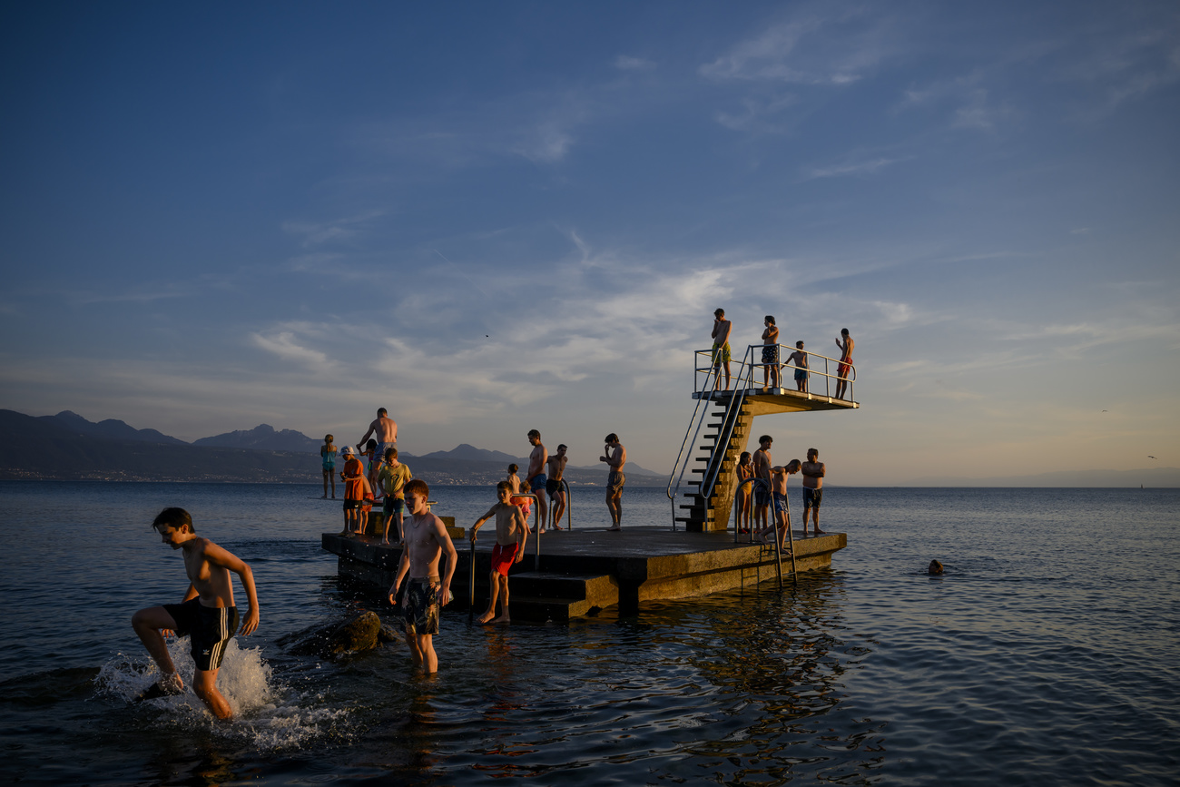 People walk in the water of Lake Leman next to a diving board during the last rays of sunshine, June 2022