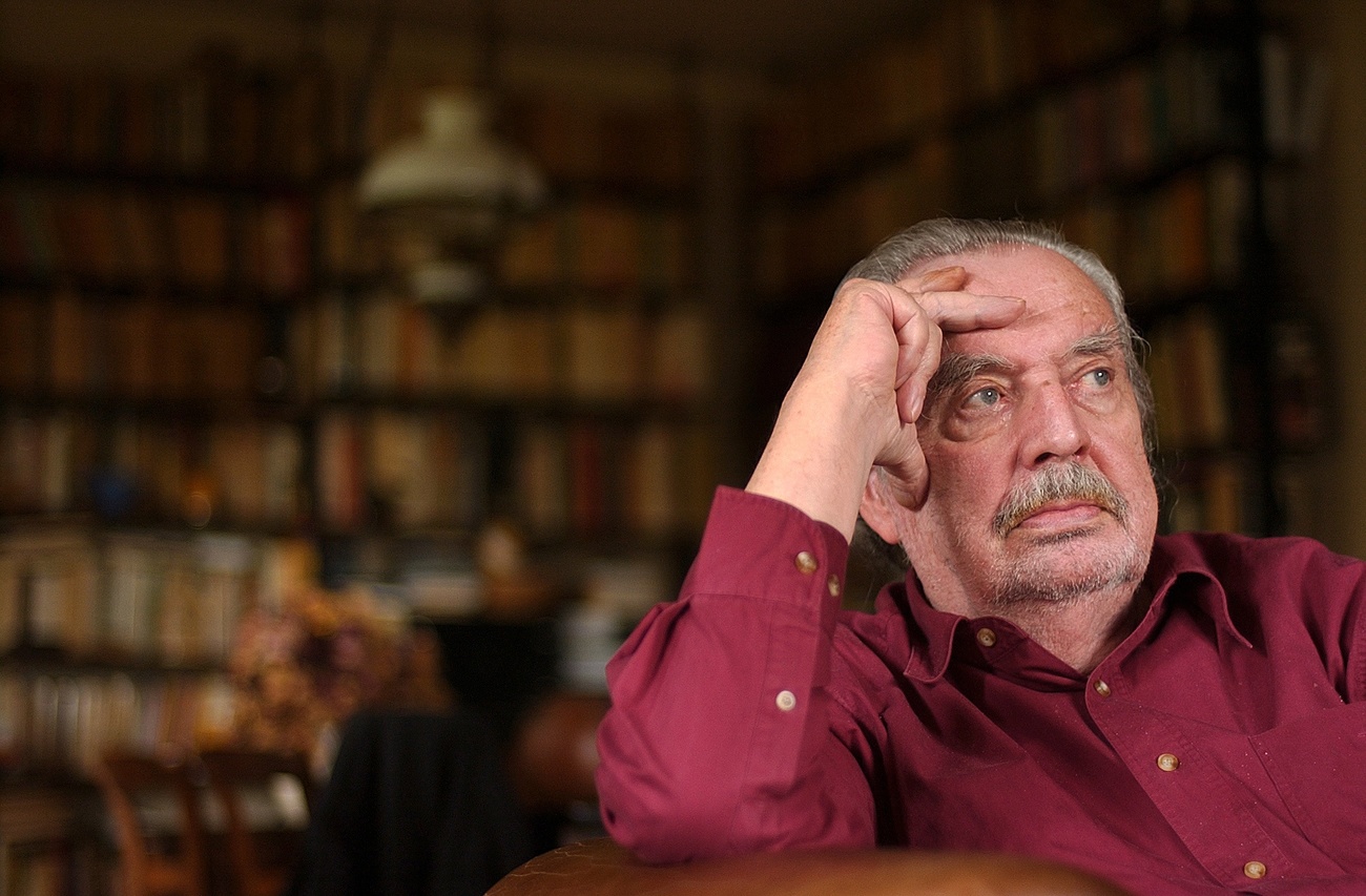 uomo anziano seduto in una biblioteca in camicia rossa appoggia la testa sulla mano destra
