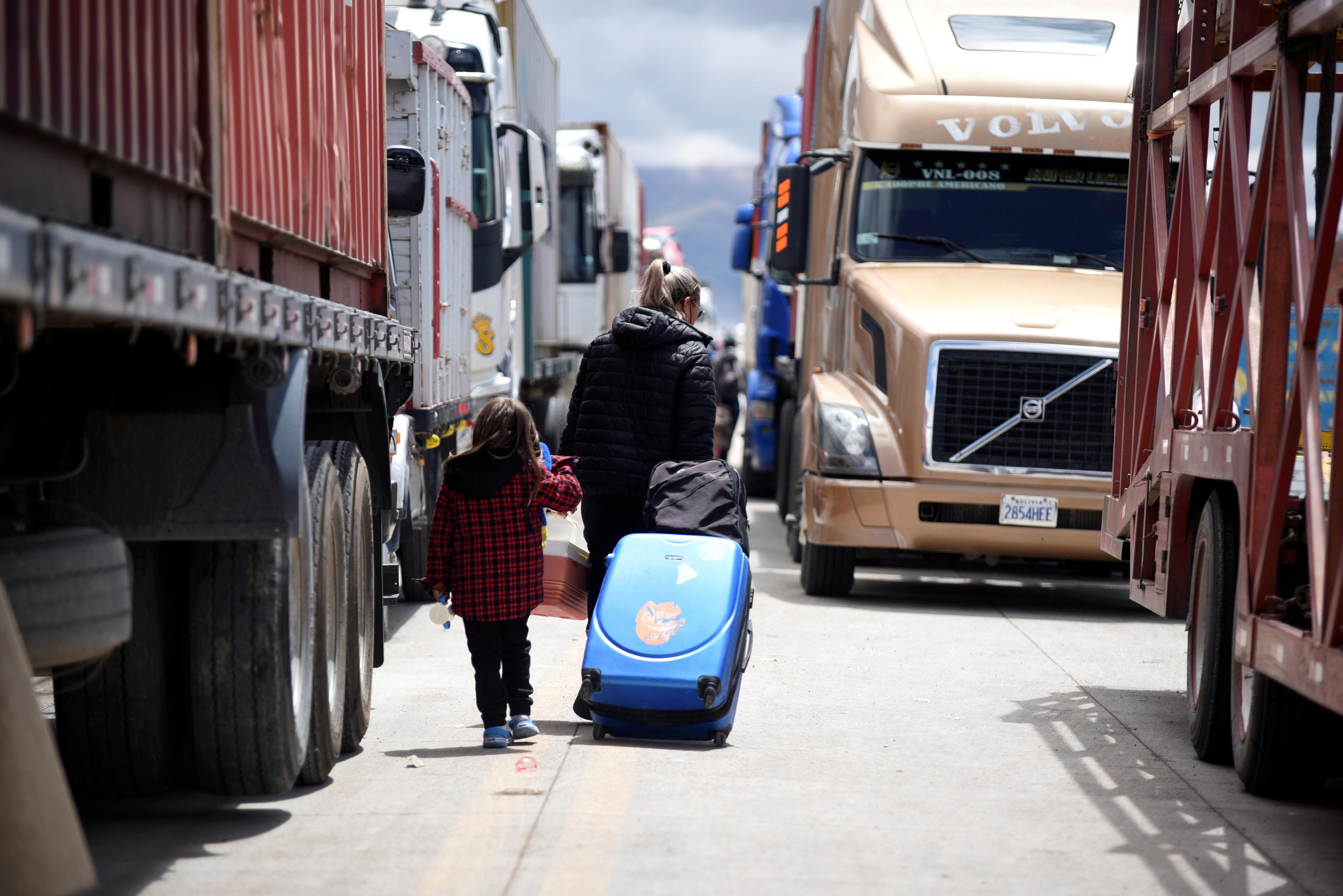 Una mujer y un menor con maletas avanzan en medio de camiones.